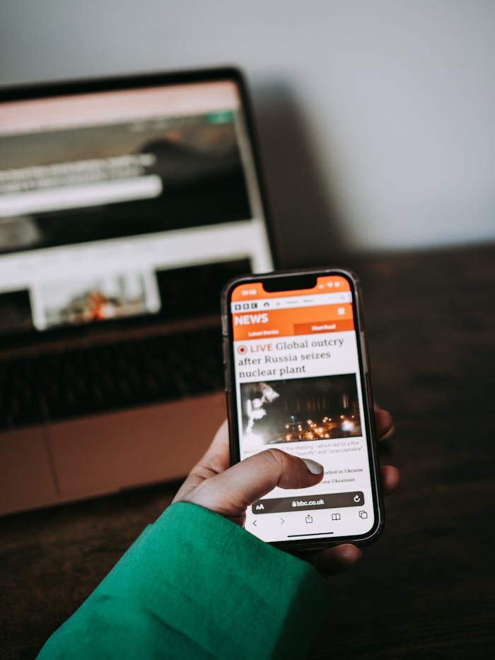 Person using smartphone for reading news online with laptop in background.